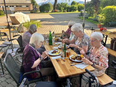 Bewohnerinnen und Bewohner sitzen an Tischen und genießen Essen und Getränke während des Grillfestes