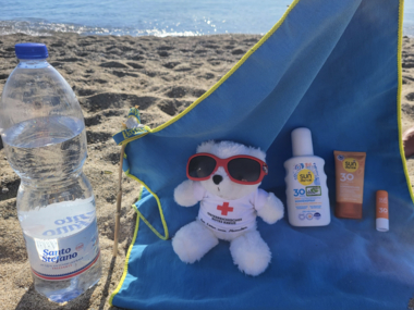 Erklärbär Bärli sitzt mit einer Sonnenbrille am Strand auf einem Handtuch im Schatten. Neben ihm befindet sich Sonnencreme und eine große Flasche Wasser.