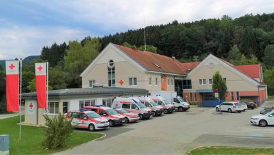 Man sieht eine Rot Kreuz Dienststelle, im Vordergrund stehen einige Autos vom RK