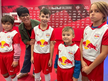 Vier junge Kinder in weiß-rotem Trikot des FC Red Bull Salzburg. Eine Dame mit Brille steht vorgebeugt im Hintergrund und umfasst die Arme des zweiten Kindes von links.