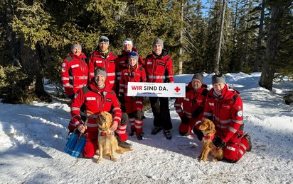 Gruppenfoto mit Mitgliedern der Suchhunde-Staffel des Roten Kreuzes Salzburg. Fünf Hunderführerinnen stehen und halten eine Tafel mit der Aufschrift "Wir sind da" in der Hand, davor knien drei Hundeführer, zwei davon mit Suchhund. Einer hält eine Lawinenschaufel in der Hand.