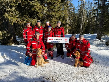 Gruppenfoto mit Mitgliedern der Suchhunde-Staffel des Roten Kreuzes Salzburg. Fünf Hunderführerinnen stehen und halten eine Tafel mit der Aufschrift "Wir sind da" in der Hand, davor knien drei Hundeführer, zwei davon mit Suchhund. Einer hält eine Lawinenschaufel in der Hand.