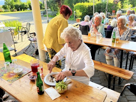 Eine Bewohnerin sitzt am Tisch und schneidet ihr Essen, im Hintergrund andere Bewohnerinnen und Bewohner