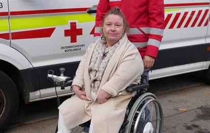 Eine Dame sitzt im Rollstuhl, hinter ihr steht ein Sanitäter in roter Uniform. Im Hintergrund ist ein Rettungswagen zu sehen.