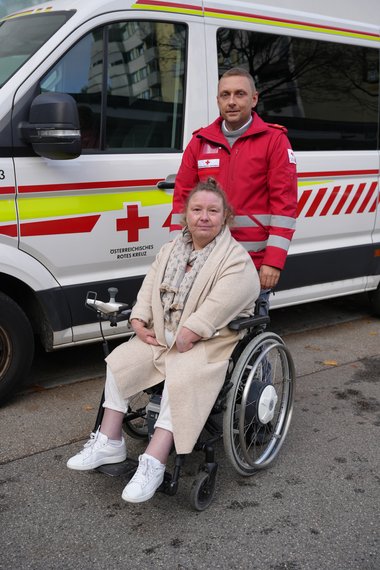 Eine Dame sitzt im Rollstuhl, hinter ihr steht ein Sanitäter in roter Uniform. Im Hintergrund ist ein Rettungswagen zu sehen.
