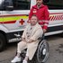 Eine Dame sitzt im Rollstuhl, hinter ihr steht ein Sanitäter in roter Uniform. Im Hintergrund ist ein Rettungswagen zu sehen.