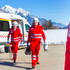 Zwei Sanitäter mit Gerätschaften gehen vom RTW Richtung eines weiteren Sanitäters, dessen Rotes Kreuz zu sehen ist.