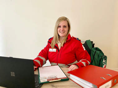 Mag. Michaela Gruber - Leiterin der Psychosozialen Betreuung - sitzt in Rotkreuz-Einsatzuniform an einem Schreibtisch. Vor ihr liegen ein Notebook, eine dicke Mappe und ein Notizblock. Auf ihrem Sessel hängt ein Rucksack mit der Aufschrift "KI - Betreuung".
