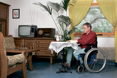 Pflege daheim - leicht(er) gemacht. Mit Pflegemittel für daheim wie z. B. mit einem Rollstuhl ist Betroffenen und ihren Familien geholfen. Am Bild sitzt ein Mann im Rollstuhl an einem Tisch und liest ein Buch.