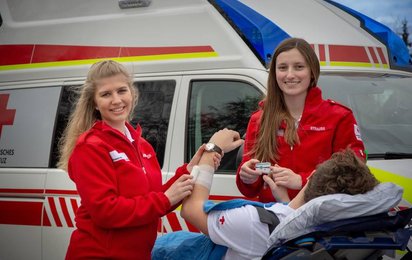 Carina und Helena sind freiwillige Sanitäterinnen - © OÖRK Zwei weibliche Sanitäterinnen vor einem Rettungsauto wie sie einen Patienten betreuen.