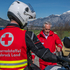 Jan Reiter, in seiner Motorrad-Einsatzuniform steht vor seinem Einsatzmotorrad und erklärt Rotkreuz-Präsident Günther Ennemoser in Rotkreuz-Uniform die Funktionsweise des Motorrades, das alles vor der Tiroler Bergkulisse