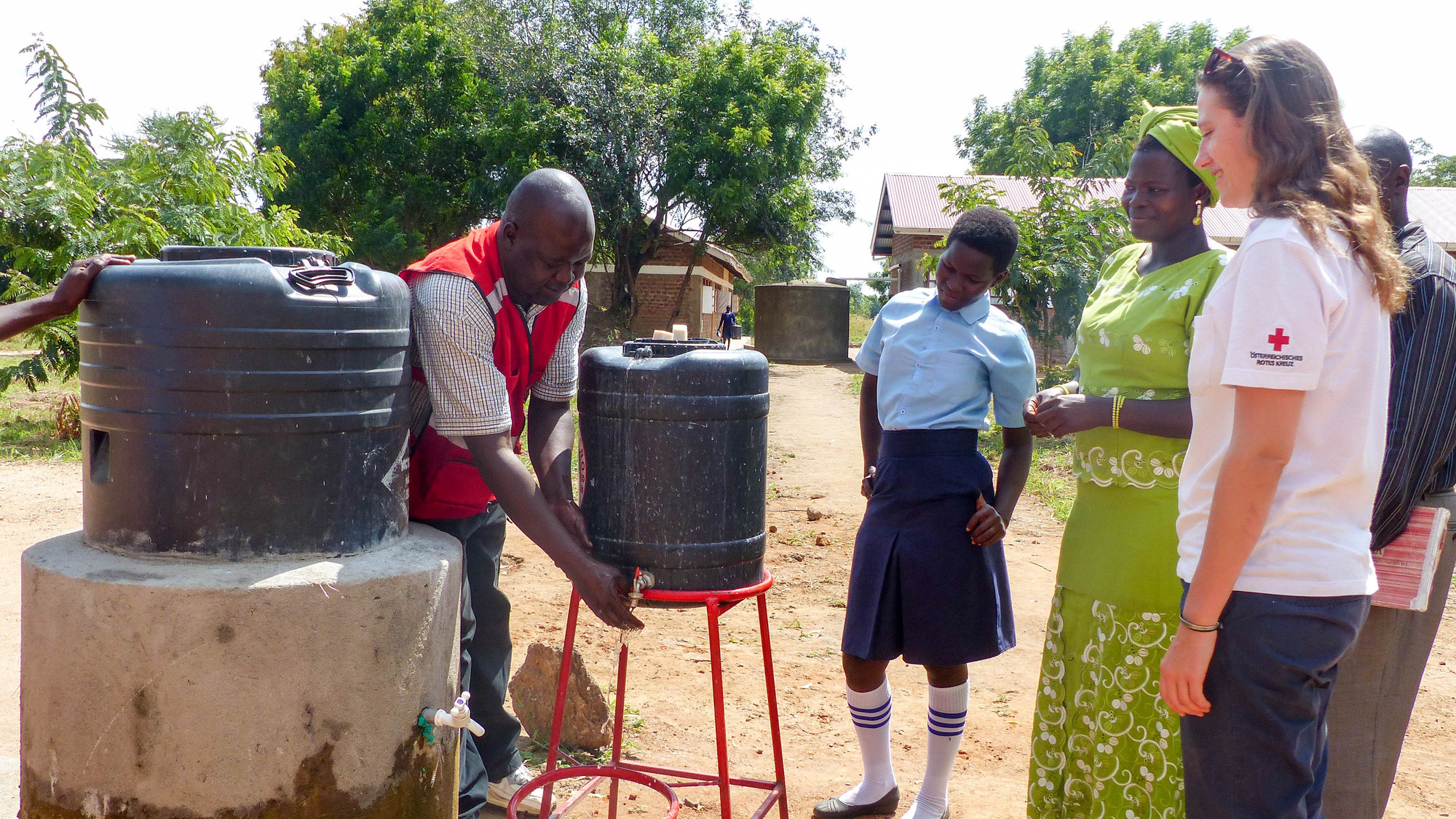 © ÖRK/Uganda Red Cross Eine Rotkreuz-Helferin in Uganda unterstützt bei einem Wasserprojekt. Ein lokaler Rotkreuz-Helfer wäscht sich die Hände unter einem Kanister, weitere Personen schauen zu.