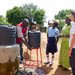 Eine Rotkreuz-Helferin in Uganda unterstützt bei einem Wasserprojekt. Ein lokaler Rotkreuz-Helfer wäscht sich die Hände unter einem Kanister, weitere Personen schauen zu.