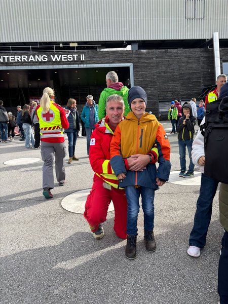 Ein Kind mit Haube und Jacke lächelt in den Kamera. Hinter ihm kniet ein Rotkreuz-Mitarbeiter und umfasst das Kind im Bereich des Bauches. Im Hintergrund das Stadion und mehrere Personen.