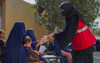 Eine Mitarbeiterin des IFRC reicht einer Frau mit einem Kind auf ihrem Arm einen Becher Wasser