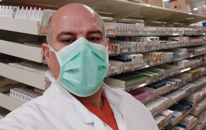 Männliche Person mit weißem Kittel und Mundschutz vor Regalen mit zahlreichen Medikamentenpackungen in einer Apotheke oder Lager