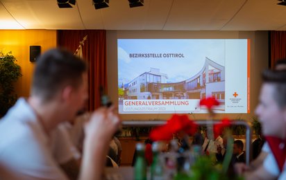 Bildschirm mit der Präsentation bei der Generalversammlung 2024. Die Gäste sitzen im Vordergrund