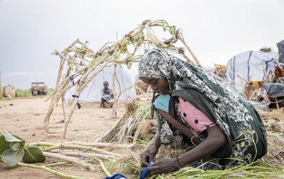 Eine sudanesische Frau sitzt in einem Flüchtlingscamp im Tschad auf dem Boden und bearbeitet mit ihren Händen Pflanzen.