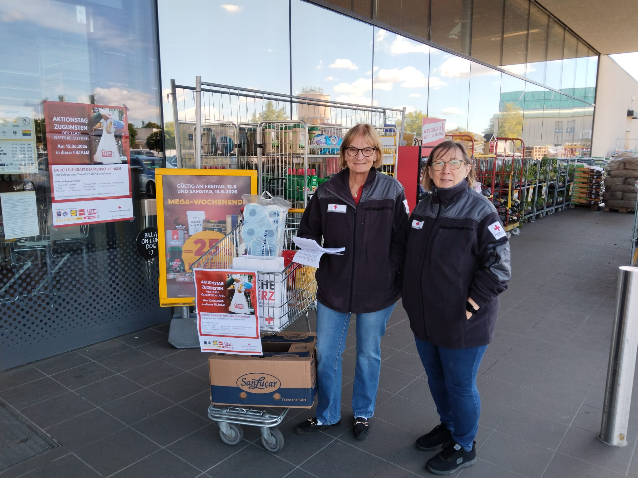 Zwei Personen in schwarzen Jacken mit Rotkreuz-Logo stehen vor einem Supermarkt neben einem Einkaufswagen mit Spendenboxen und Informationsplakaten.