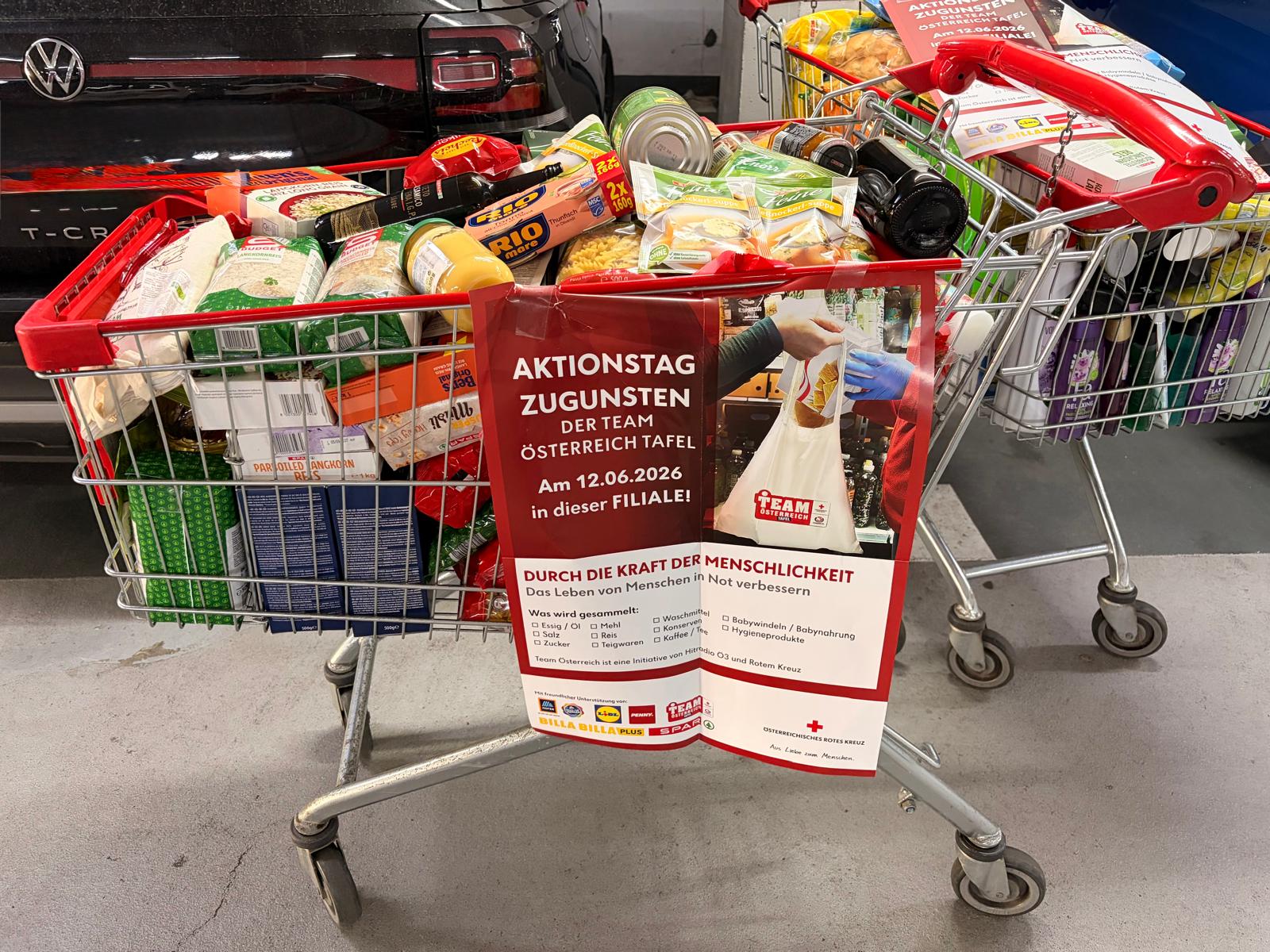 Einkaufswagen mit haltbaren Lebensmitteln und Hygieneartikeln sowie einem roten Plakat mit der Aufschrift 'AKTIONSTAG ZUGUNSTEN DER TEAM ÖSTERREICH TAFEL Am 12.06.2026 in dieser FILIALE!'