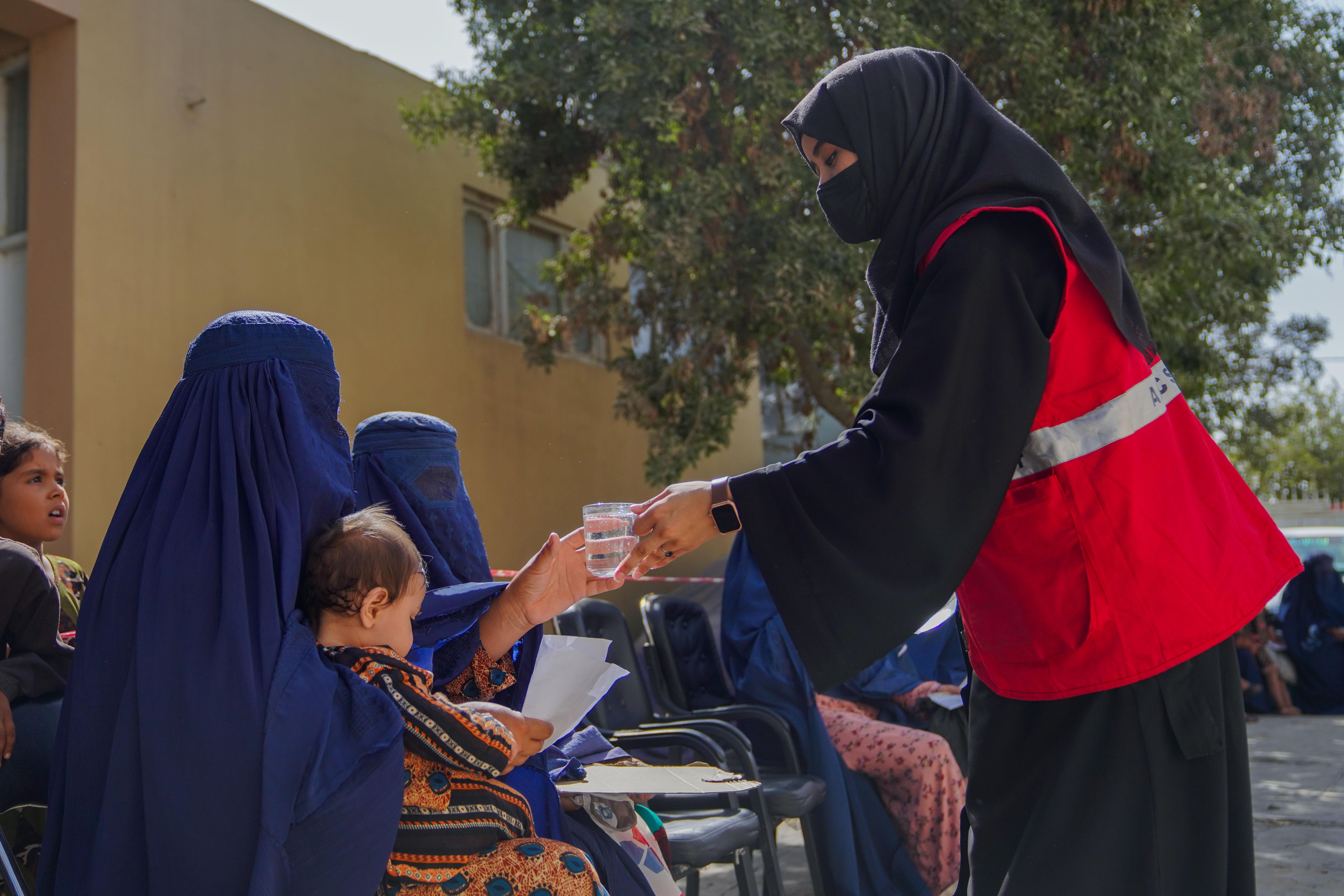 Eine Mitarbeiterin des IFRC reicht einer Frau mit einem Kind auf ihrem Arm einen Becher Wasser