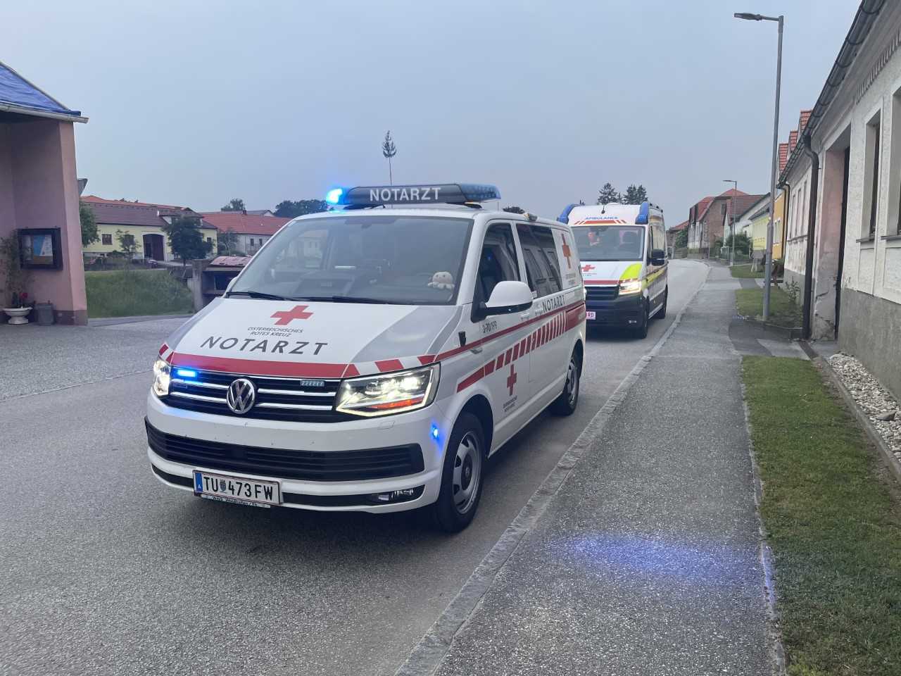 Ein Notarzteinsatzfahrzeug und ein Rettungstransportwagen im Einsatz auf einer Straße im ländlichen bewohnten Bereich.