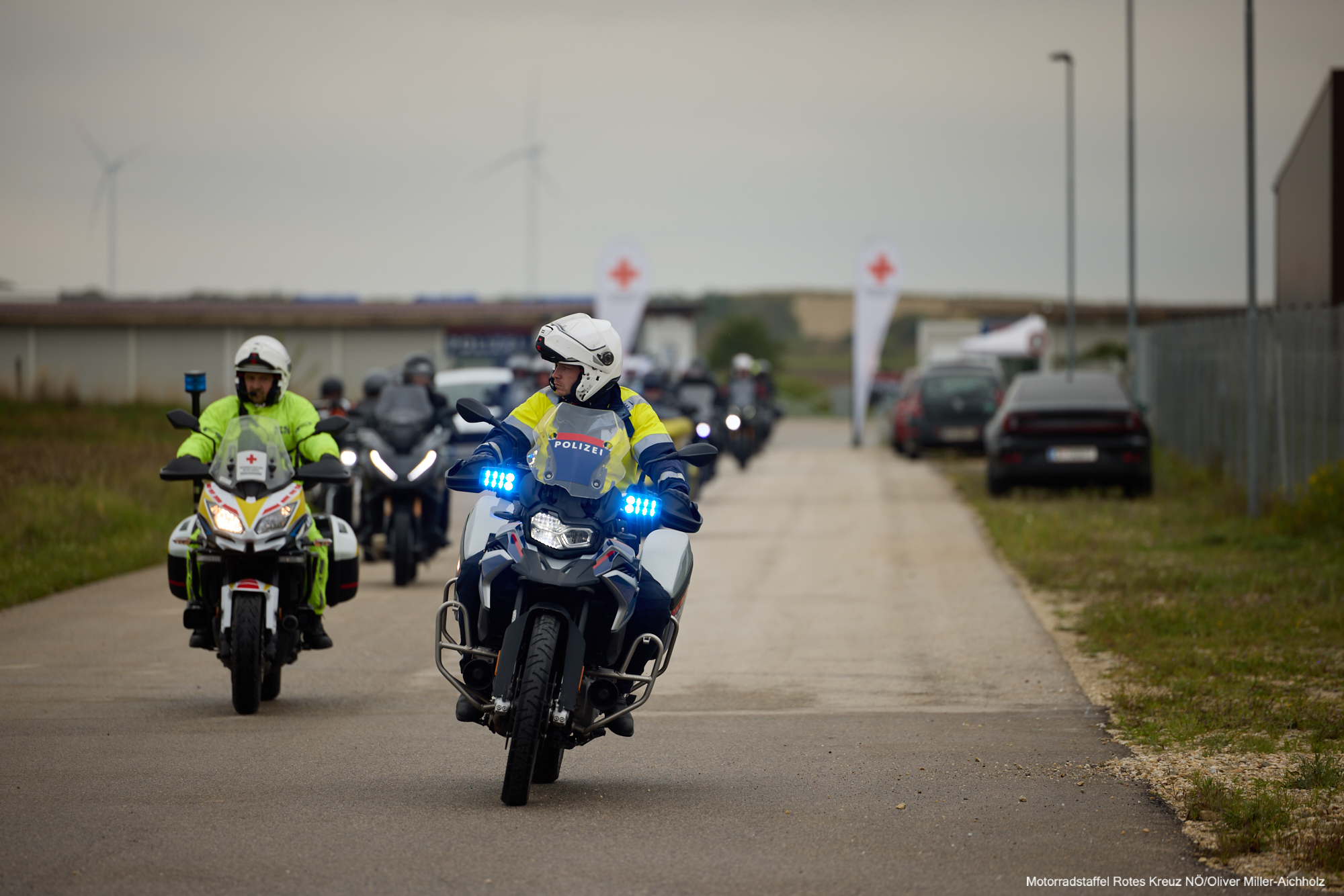 Ein Rotkreuz-Motorrad und ein Polizei-Motorrad fahren auf das Gelände zu - dahinter sieht man die anderen Motorräder.