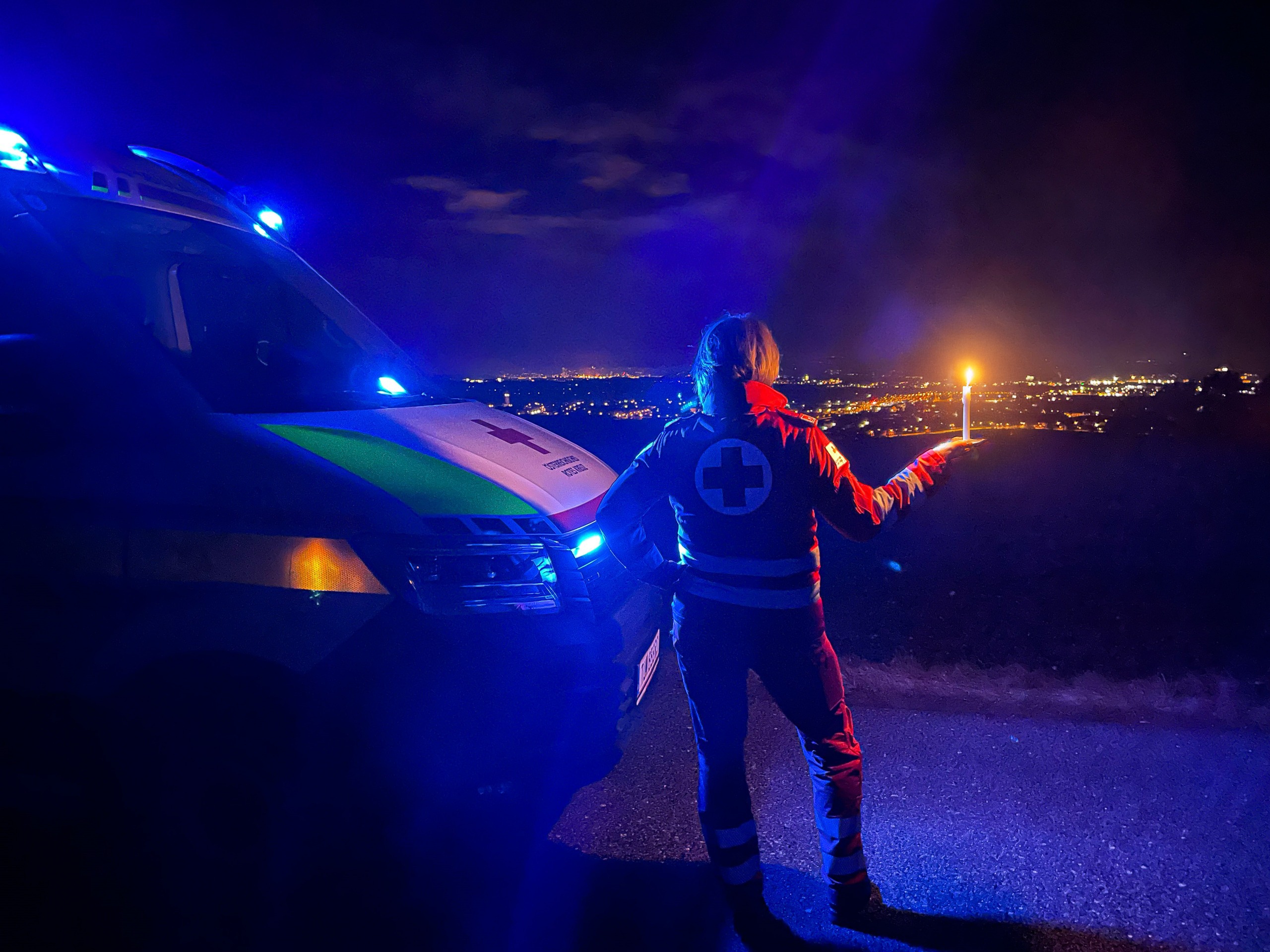 Eine Rotkreuz-Mitarbeiterin steht mit einer Kerze vor einem Rettungsauto, es ist dunkel und sie blickt auf eine beleuchtete Stadt hinunter.