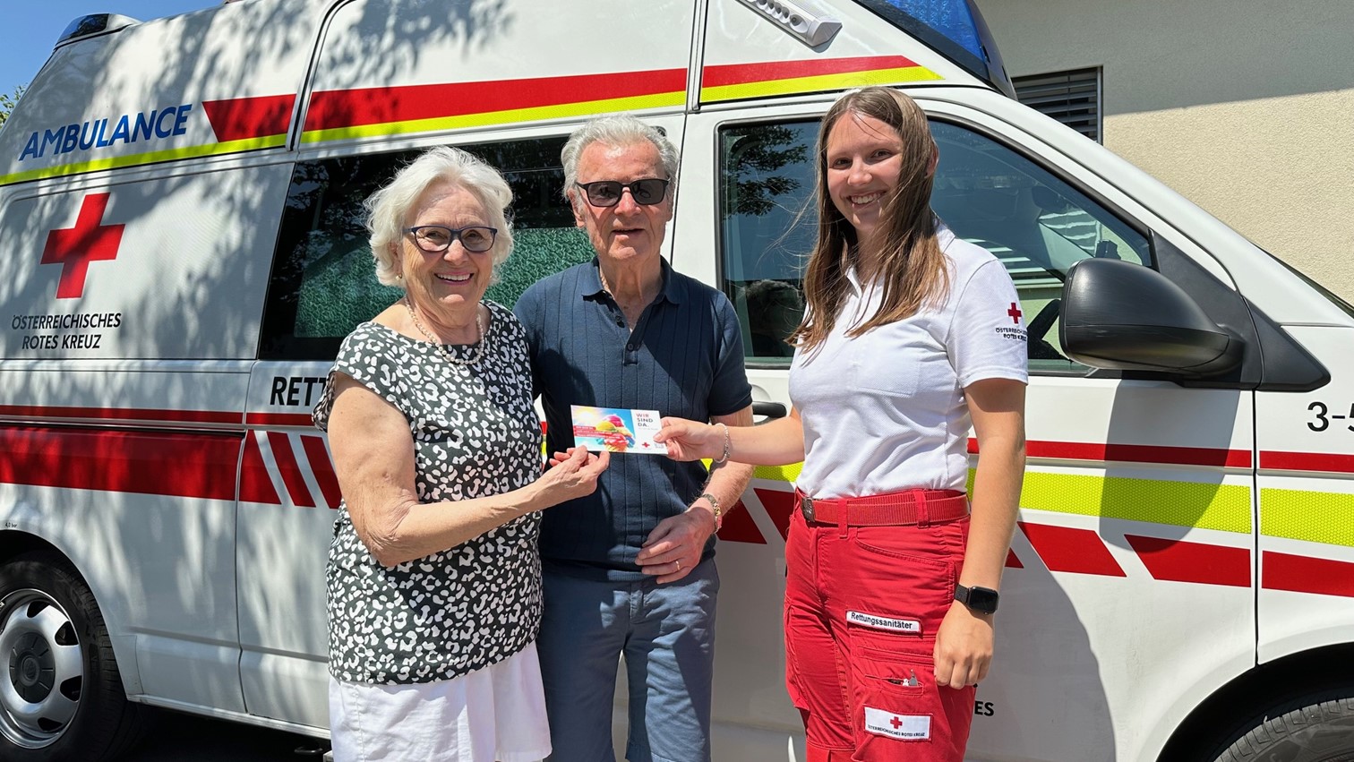 Eine Rotkreuz-Mitarbeiterin überreicht eine Hitzebuddy-Freecard älternen Menschn vor einem Rettungsauto.