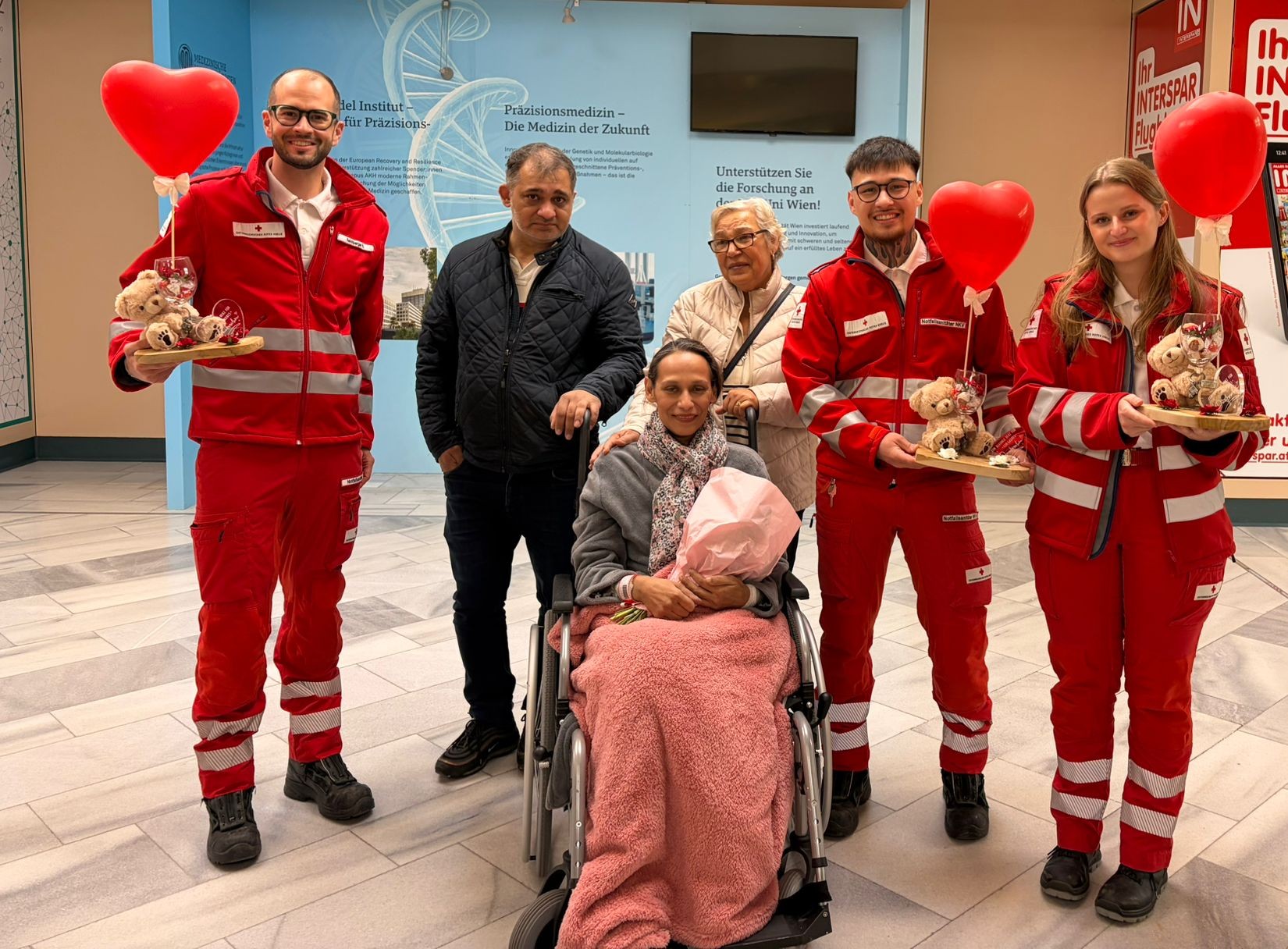 Rotkreuz-Mitarbeiter:innen mit der Patient:innen im Rollstuhl, sie hält einen Blumenstrauß in der Hand.