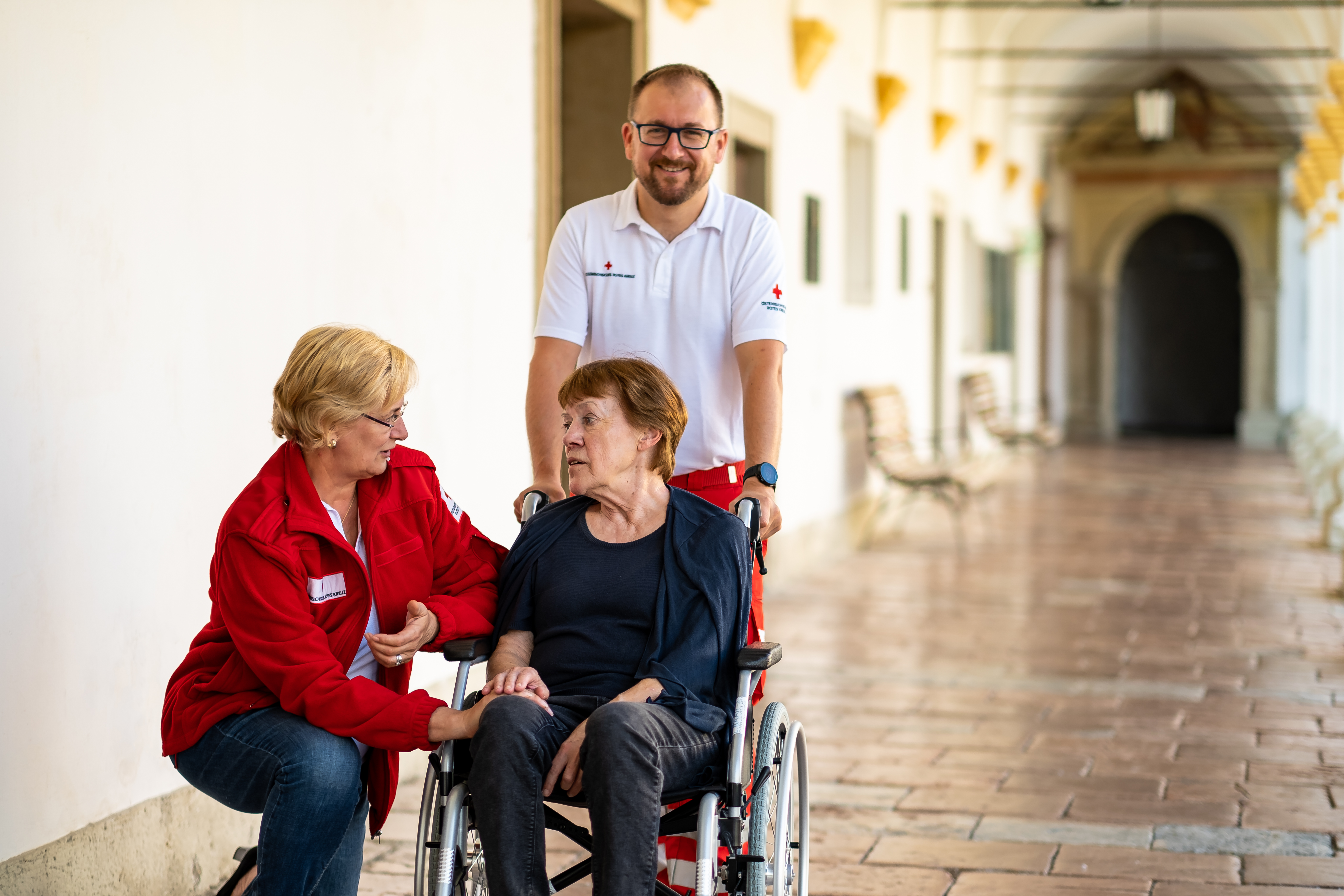 Eine Frau im Rollstuhl wird von zwei RK-Mitarbeitern betreut