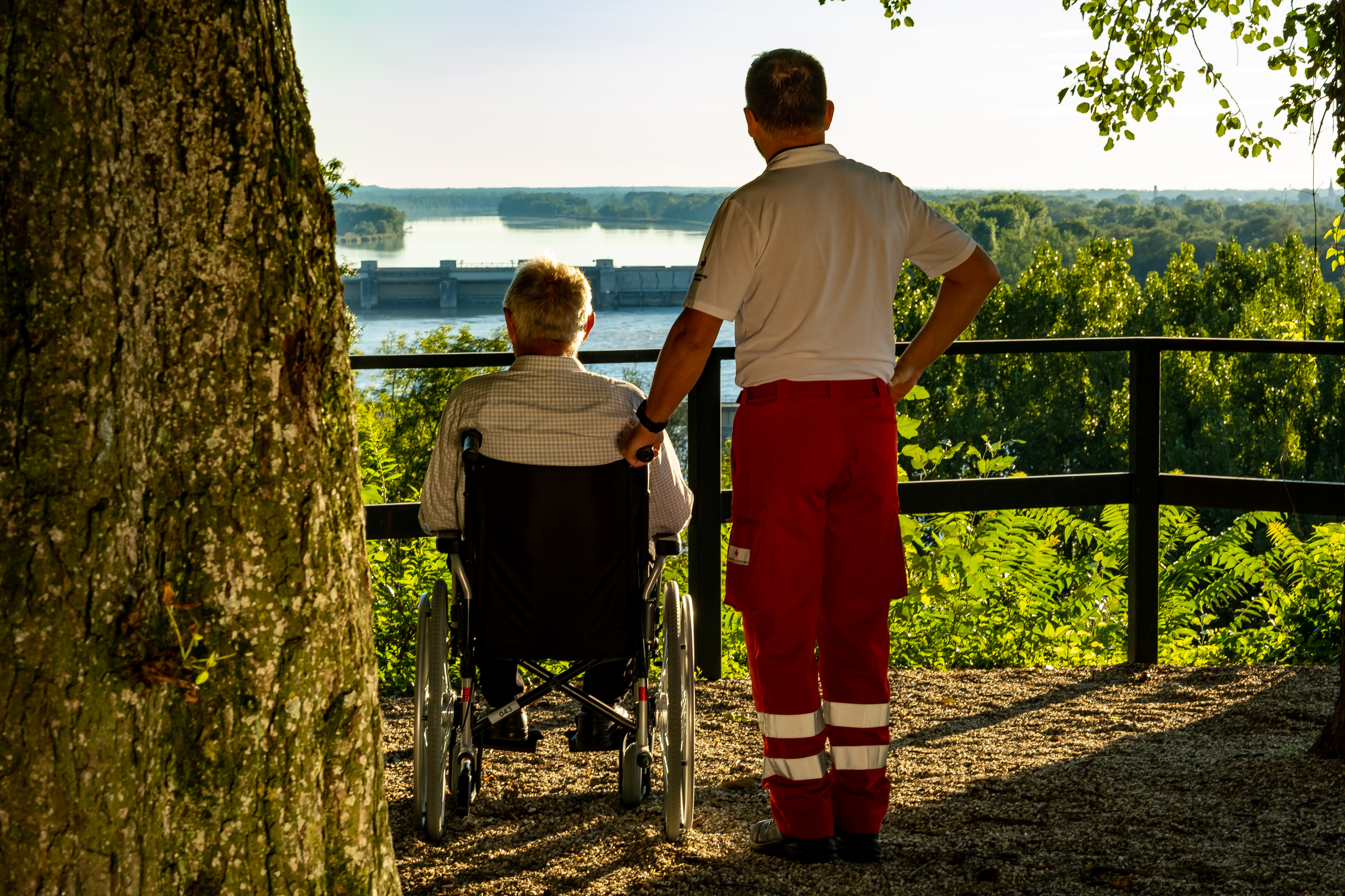 älterer Herr im Rollstuhl steht auf einer Aussichtsplattform, neben ihm ein Rotkreuz-Mitarbeiter