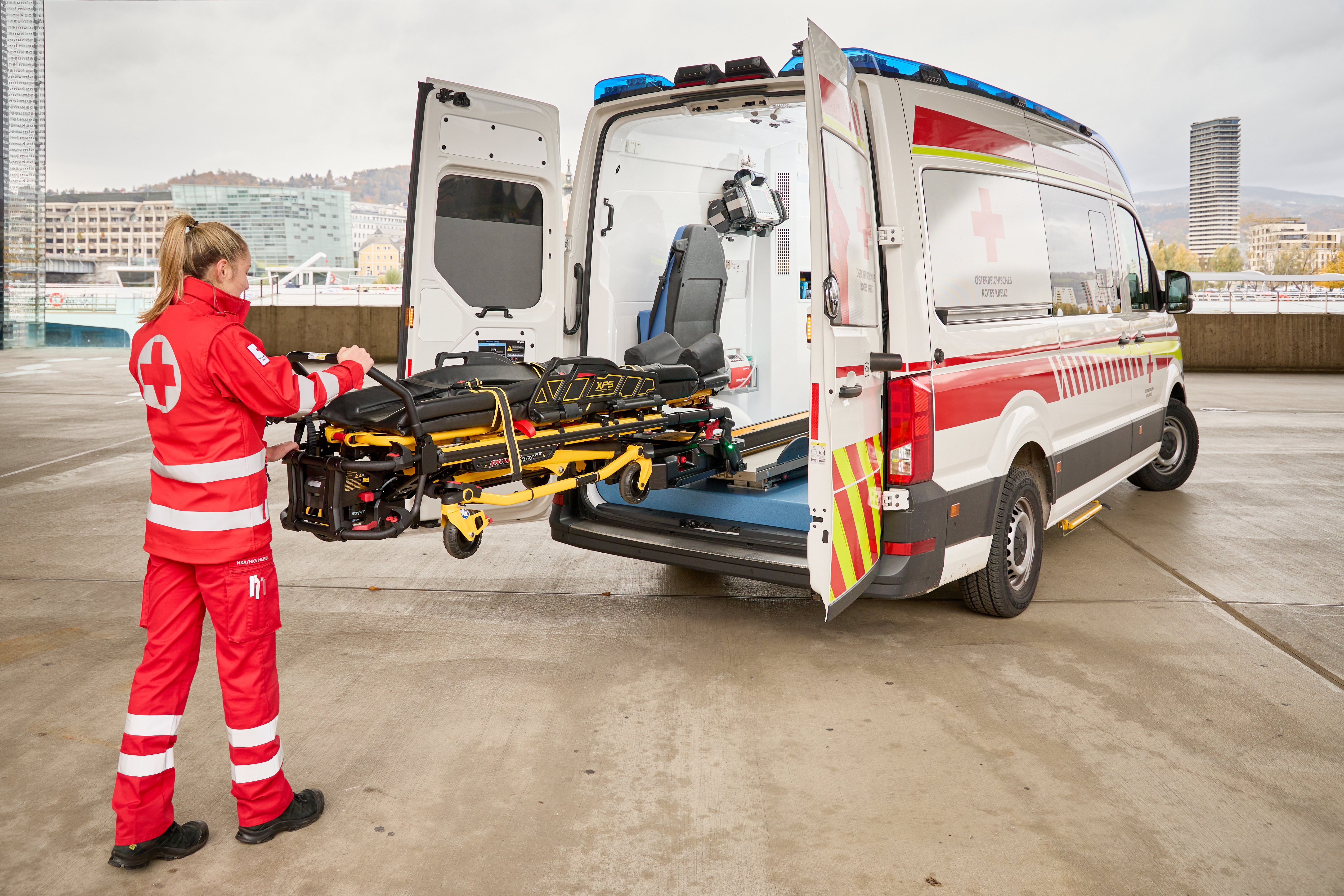 Eine Rettungssanitäterin steht hinten am Auto und zeigt die voll elektische Fahrtrage des Rettungswagens.