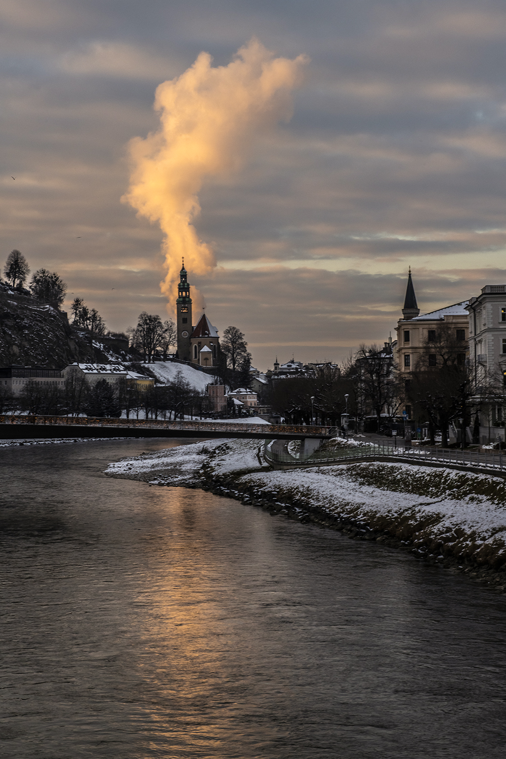 Blick über die Salzach nach Mülln. Eine Wolke scheint aus dem Kirchtum der Müllner Kirche aufzusteigen.