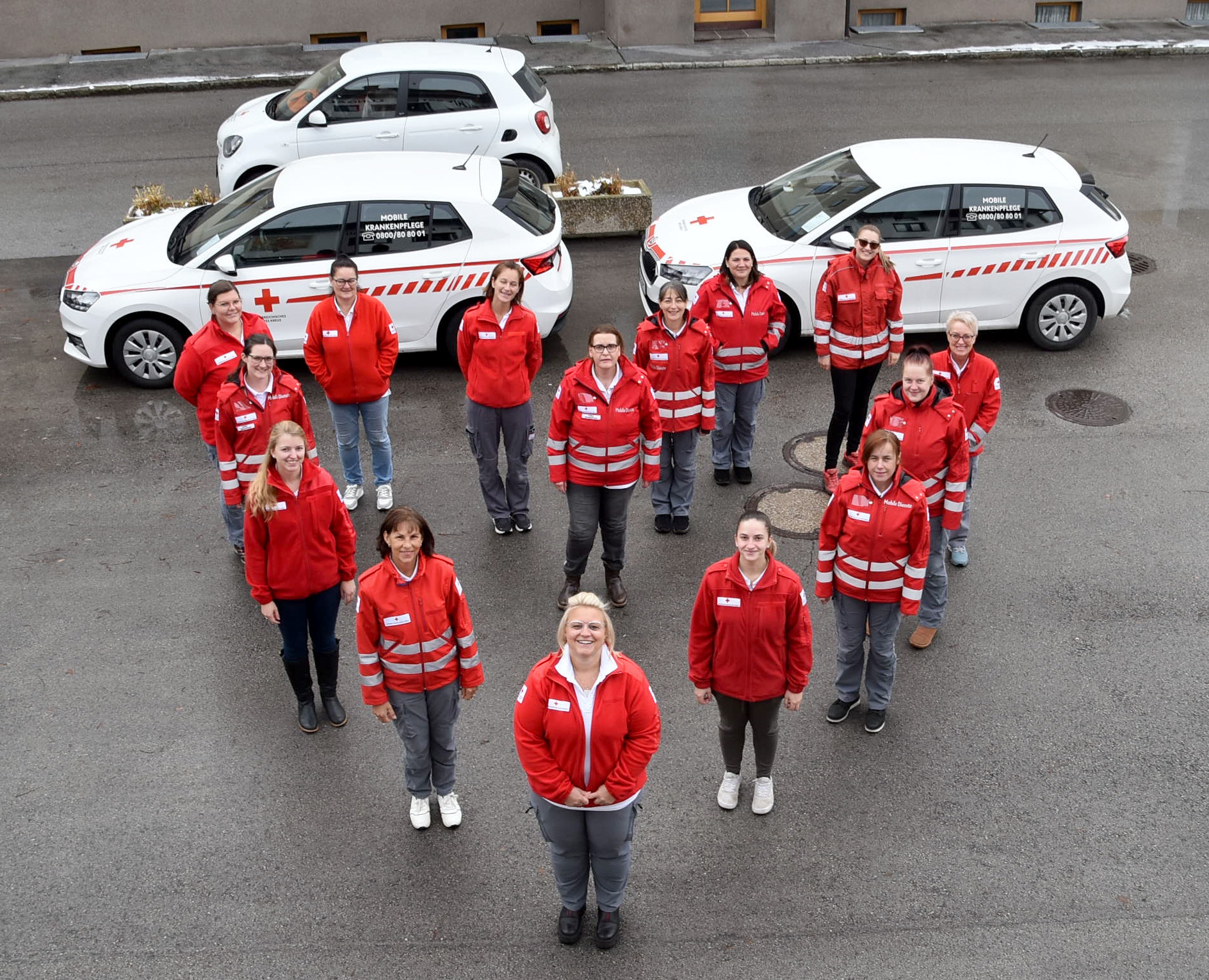 Mitarbeiterinnen der Mobilen Krankenpflege stehen in Form eines Herzens vor Rotkreuz-Fahrzeugen