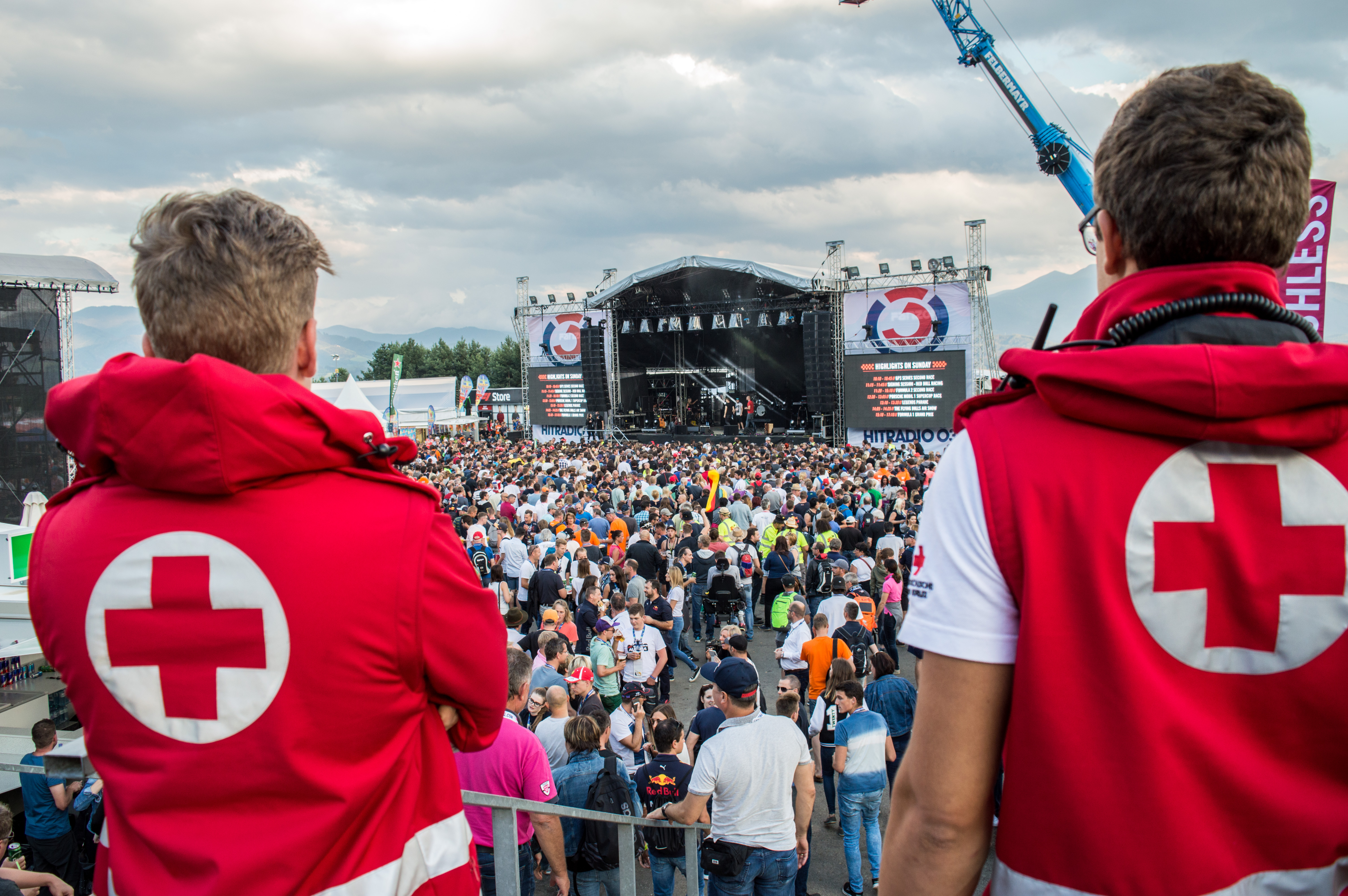Man sieht zwei Sanitäter von hinten, die die Menschenmenge eines Konzertes überblicken. Das Konzert findet im Freien statt - es ist bewölkt.