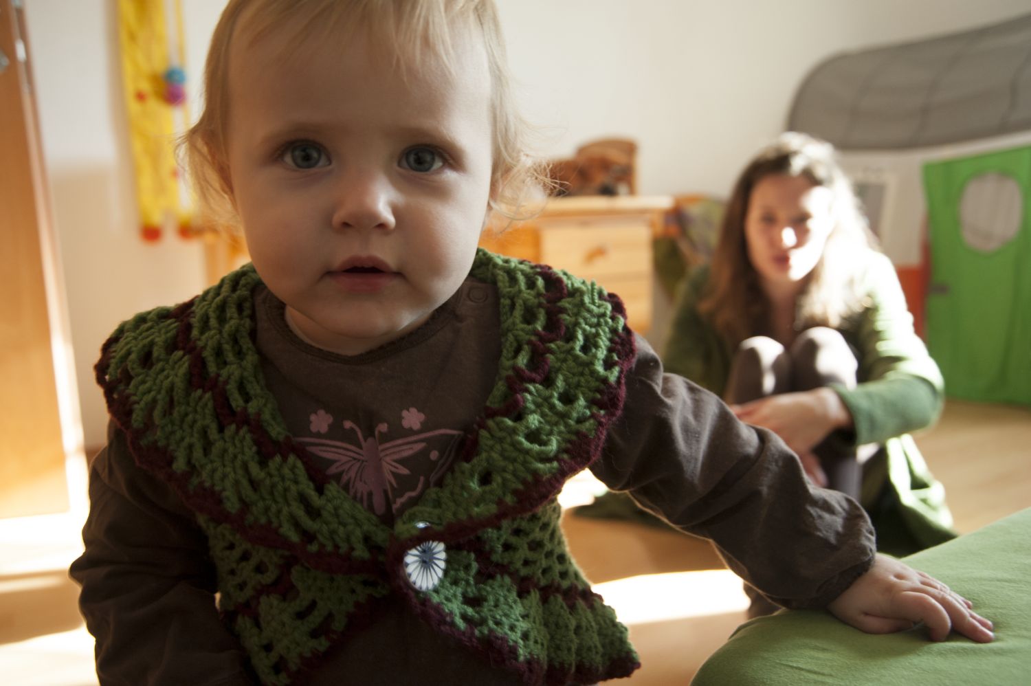 im Vordergrund krabbelt ein Kleinkind, im Hintergrund sieht man die Mutter, die am Boden sitzt und das Kind beobachtet